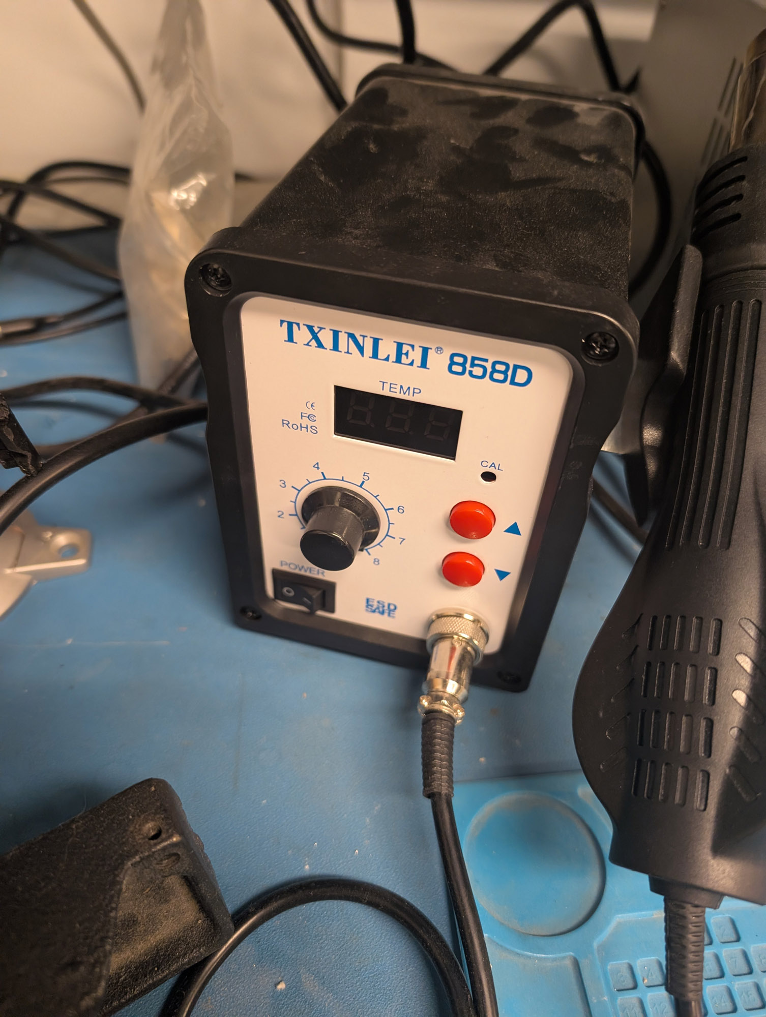 A hot air reflow station that says "TXINLEI 858D" at the top, with a three digit TEMP display below that. Thereis a 0-8 knob under that, as well as up and down arrow buttons on the right side. A power rocker switch is at the bottom, and the cable connected to the handpiece which is in a holder on the right side, with a fan built into the base of the handpiece